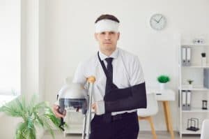 Man in business attire with arm in a sling, crutch, and bandaged head holding a motorcycle helmet, standing in a modern office environment.