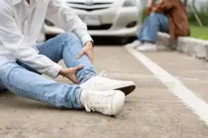 Person sitting on the road holding their ankle in apparent pain, with a car and another person visible in the background.