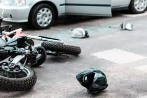 A crashed motorcycle lies on the ground near a helmet and shattered car parts, next to a parked car with an open door and broken window.
