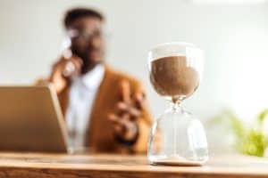 A glass hourglass filled with sand sits on a desk in the foreground; in the background, a person speaks on the phone next to a laptop.