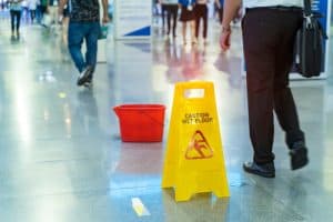 A yellow "Caution Wet Floor" sign and a red bucket are placed on a shiny indoor floor as people walk by.