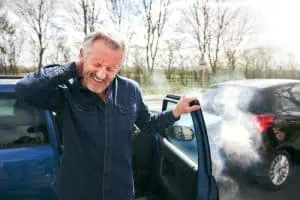 Man standing beside open car door, holding his neck in pain, with smoke coming from the vehicle after a car accident.