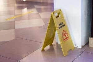 A yellow caution sign warning of a wet floor stands on a tiled indoor surface near a white column.