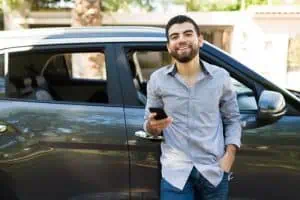 A man in a gray shirt leans against a dark-colored car, holding a smartphone and smiling, with trees and buildings in the background.