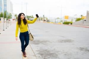 Woman with curly hair wearing a yellow sweater holds up her phone while walking on a sidewalk in an urban area.