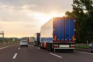 Several trucks and a car drive on a multi-lane highway surrounded by trees under a partly cloudy sky at sunset.