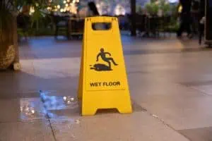 A yellow "Wet Floor" caution sign is placed on a tiled floor next to a visible puddle of water in an indoor public area.
