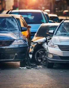 Three cars involved in a collision on a city street, with visible damage to bumpers and windshields under daylight.