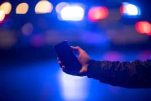 A person holds a smartphone in their hand with blurred blue and red police lights in the background at night.