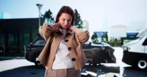 A woman stands in front of two crashed cars, holding her neck with a pained expression, suggesting she may be injured after a car accident.
