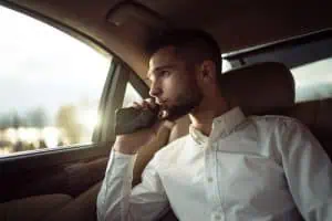 A man in a white shirt sits in a car, looking out the window while holding an electronic vape device to his mouth.