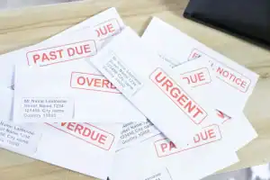 A pile of white envelopes on a table, each labeled with terms like "PAST DUE," "URGENT," "OVERDUE," and "FINAL NOTICE" in bold red letters.