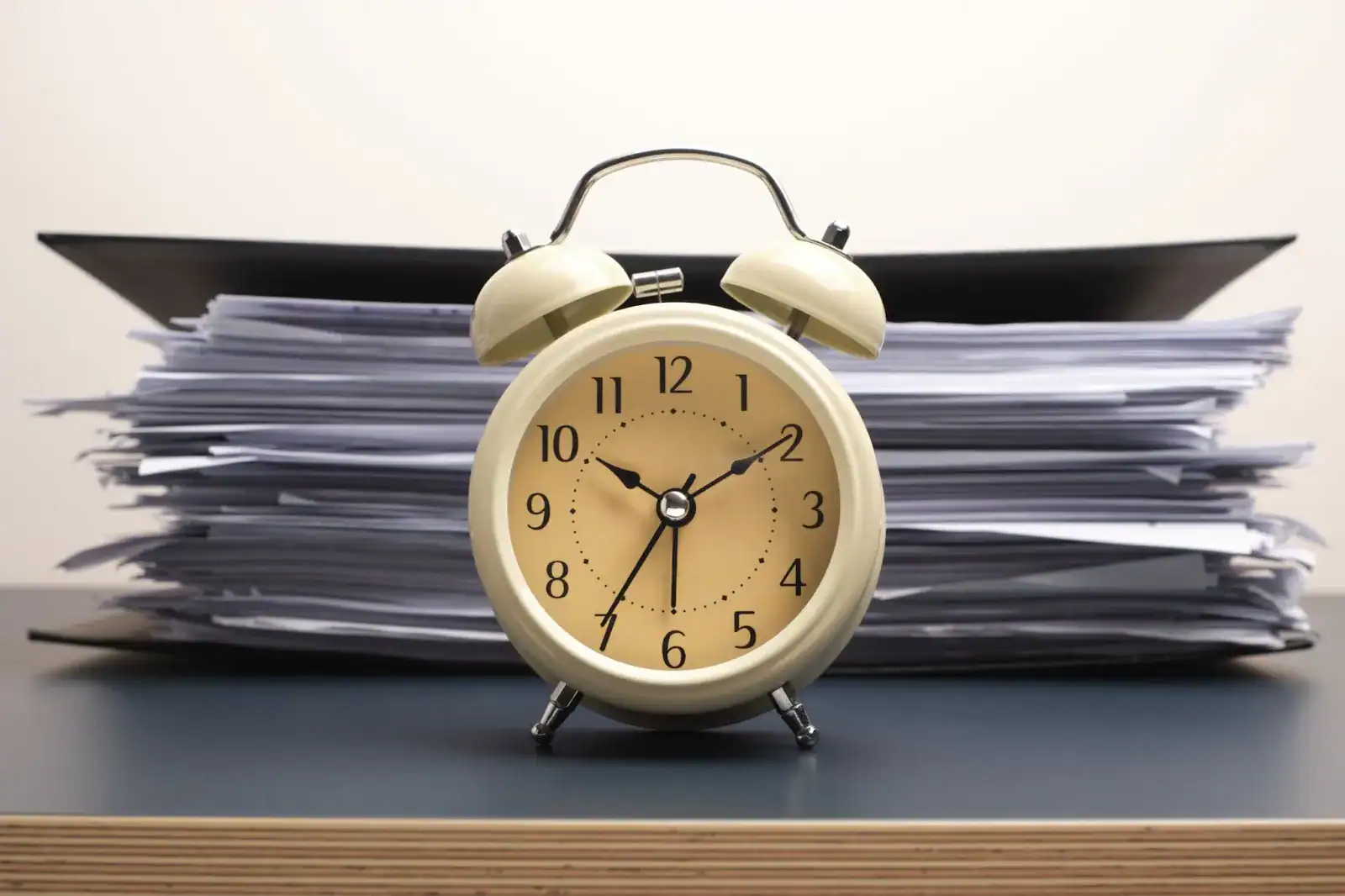 A beige alarm clock is placed in front of a large stack of documents and folders on a desk.