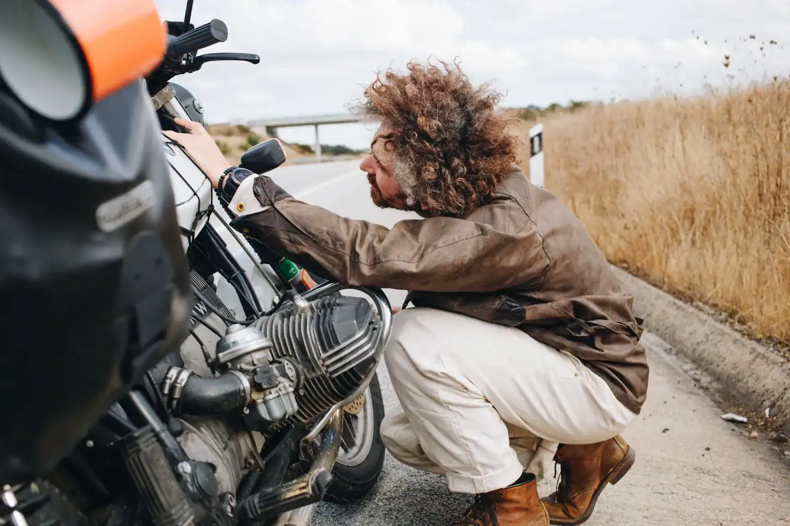 A person in a brown jacket and boots is crouched beside a motorcycle on the roadside, examining the engine. Dry grass and an empty road are in the background.