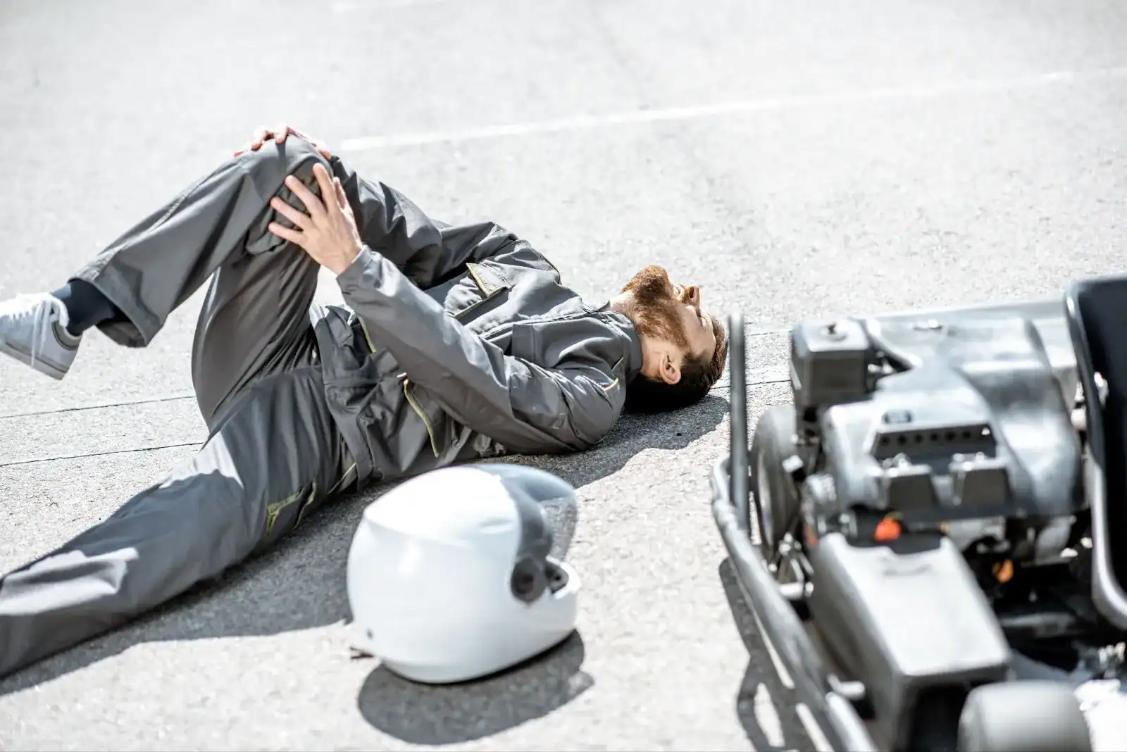 A person in a gray suit lies on the ground holding their knee next to a helmet and a go-kart, suggesting a possible crash or injury.