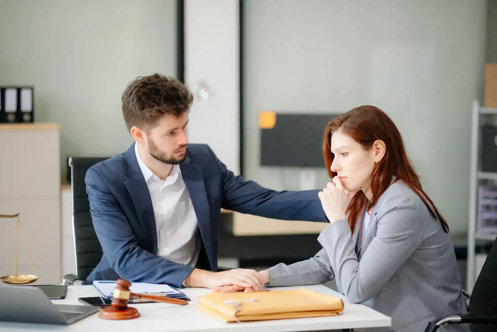 A man in a suit consoles a thoughtful woman in an office setting with legal documents, a gavel, and scales of justice on the table.