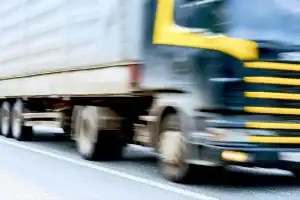 A large truck with a yellow and black cab is moving quickly along a paved road, with the image showing motion blur.