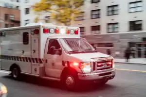 An ambulance with flashing lights drives down a city street past blurred buildings and trees during the day.