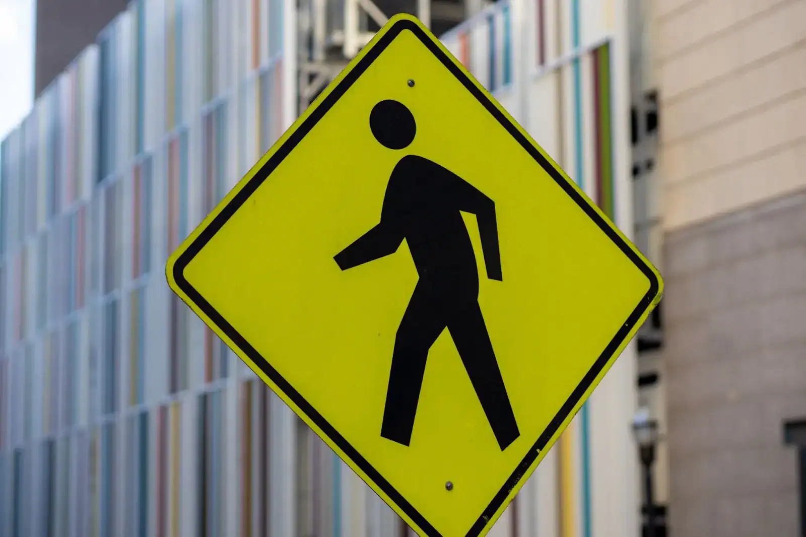 Yellow pedestrian crossing sign with a black walking figure in front of a building with vertical colorful lines.
