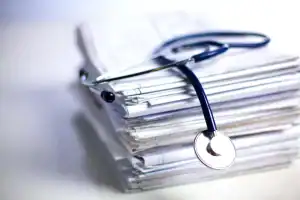 A stethoscope rests on top of a large stack of medical documents and files on a white surface.