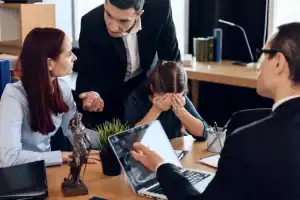 A man speaks to a woman while a child sits between them covering her face; another man with a laptop sits nearby at a desk with legal documents and a Lady Justice statue.