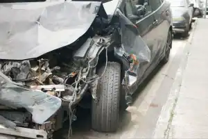 A black car with severe front and side damage is parked on the street, showing exposed internal components and a bent wheel.