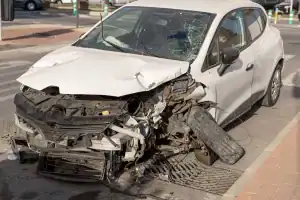 A white car with severe front-end damage and a shattered windshield is parked on the side of a street after a crash.