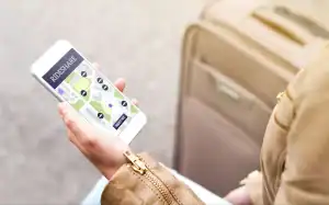 Person holding a smartphone with a rideshare app open, showing a map and ride options, next to a beige suitcase.