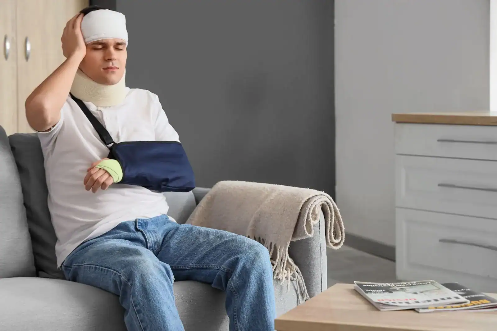 A man with a bandaged head, neck brace, and arm sling sits on a sofa, appearing to be in pain and holding his head with one hand.