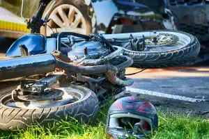 A motorcycle lies on its side near a helmet on grass after an apparent road accident, with damaged cars in the background.