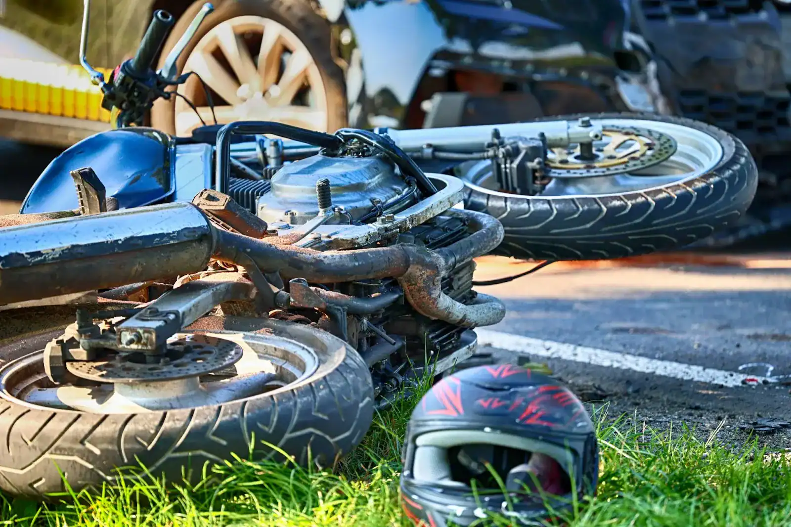 A motorcycle lies on its side near a helmet on grass after an apparent road accident, with damaged cars in the background.
