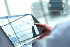 A person uses a digital touchscreen displaying a trucking log with line graphs and schedule details; trucks are visible in the blurred background.