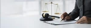 A person in a suit signs a document at a desk with a gavel and scales of justice placed nearby.