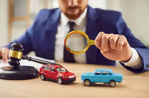A person in a suit examines model cars with a magnifying glass next to a gavel on a desk, suggesting a legal or insurance context.