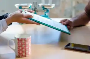 Two people sit at a table; one hand passes a clipboard with documents to the other. A coffee mug, a smartphone, and blue weighing scales are also on the table.