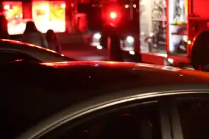 Emergency vehicles with flashing lights are parked at night. Two people are visible in the background near the illuminated vehicles.