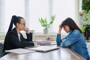 Two women sit across from each other at a desk, appearing to have a serious conversation; one woman looks concerned with her head in her hand.