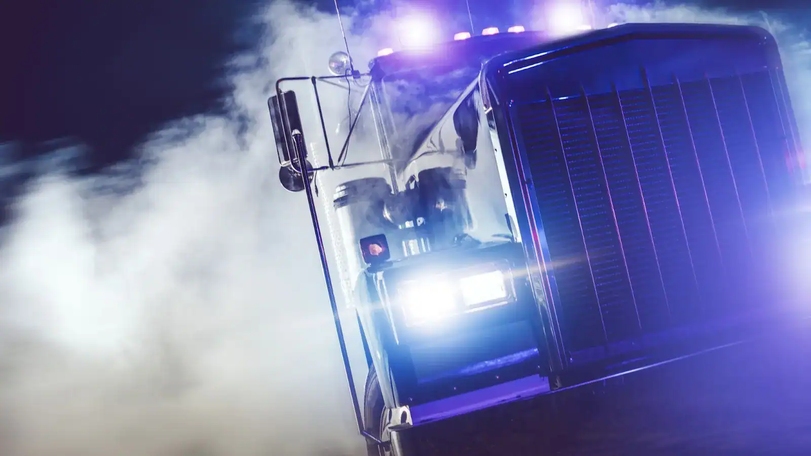 A large truck with headlights on drives through dense smoke at night, illuminated by blue and purple lights.