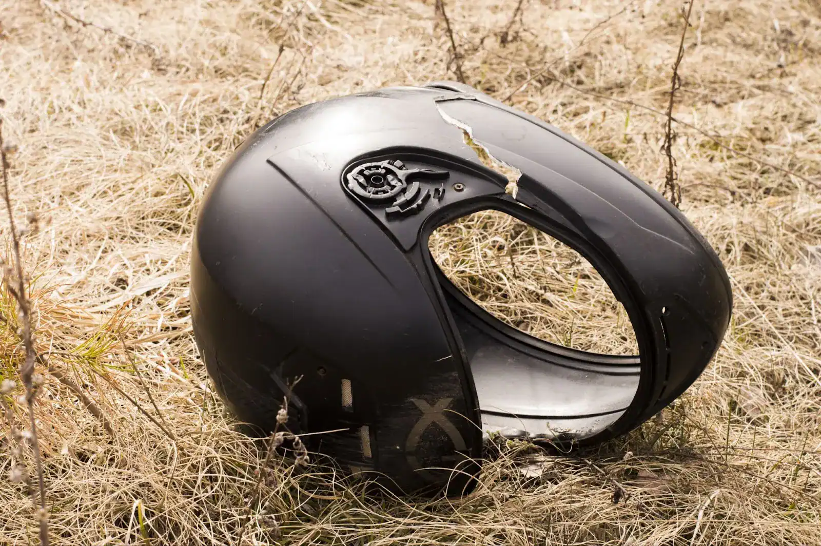 A cracked and damaged black motorcycle helmet lies on dry, yellow grass.
