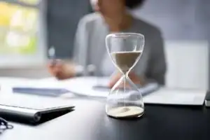 An hourglass with sand flowing sits on a desk in the foreground, while a person works with documents in the blurred background.