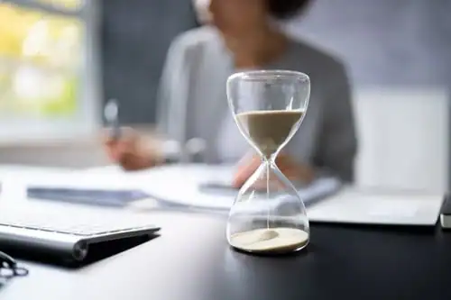 An hourglass with sand flowing sits on a desk in the foreground, while a person works with documents in the blurred background.