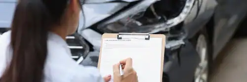 A person holding a clipboard inspects and takes notes in front of a damaged car.