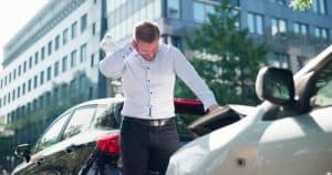 A man stands between two cars involved in a minor accident, holding his neck and looking distressed.