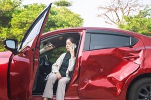 A woman sits in a red car with a large dent on the side, holding a phone to her ear with the door open.