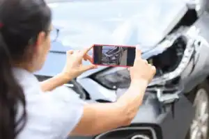 A woman takes a photo of the damaged front end of a car after an accident.