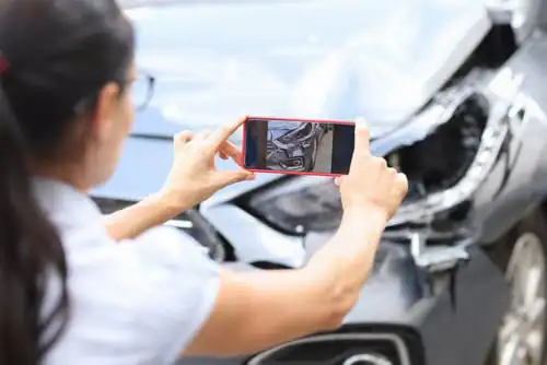 A woman takes a photo of the damaged front end of a car after an accident.