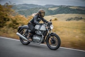 A person wearing a helmet and leather jacket rides a motorcycle on a road through a hilly, scenic landscape.