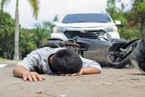 A person lies face down on the road in front of a fallen motorcycle and a white car, suggesting a traffic accident scene.