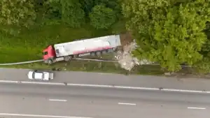 Aerial view of a red semi-truck off the road near trees, with debris spilled beside it and a white vehicle parked nearby on the roadside.
