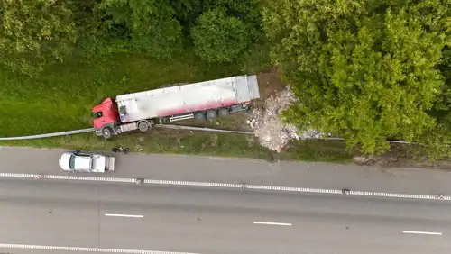 Aerial view of a red semi-truck off the road near trees, with debris spilled beside it and a white vehicle parked nearby on the roadside.